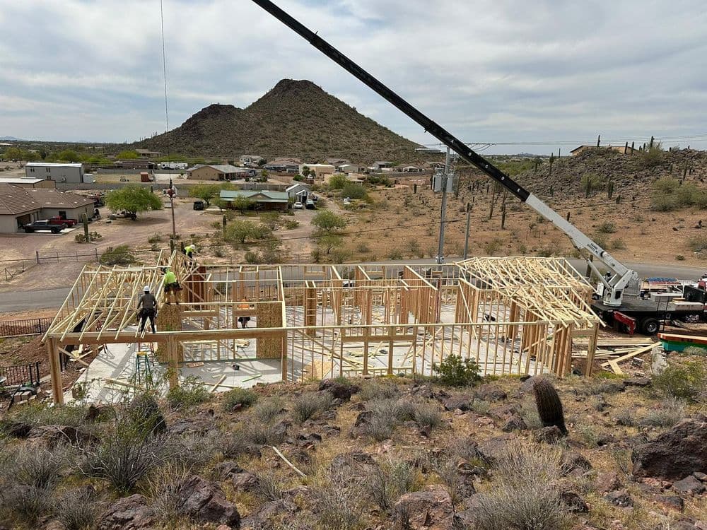 Construction of a timber-framed home with workers and machinery in a desert landscape.